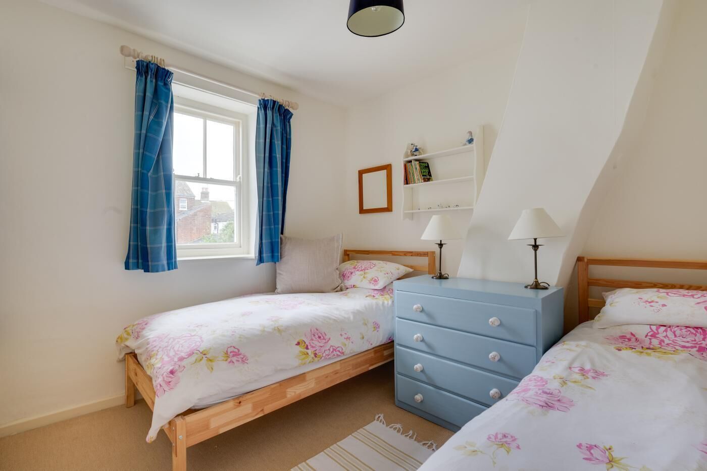 A bedroom with two single beds, a dresser, a window with curtains, a shelf with books, and a lamp.