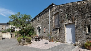 Un bâtiment en pierre avec une porte blanche, une petite fenêtre et une cour dallée avec une table et des chaises.