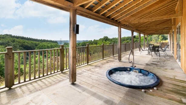 Une terrasse en bois avec un jacuzzi, du mobilier d’extérieur et une vue panoramique sur une zone boisée.
