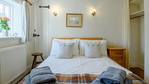 Ein Schlafzimmer mit einem Bett, einem kleinen Hocker, einem Fenster mit Vorhängen, einem Heizkörper und einem an der Wand hängenden Bild.