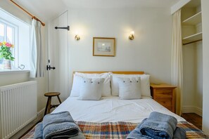 Ein Schlafzimmer mit einem Bett, einem kleinen Hocker, einem Fenster mit Vorhängen, einem Heizkörper und einem an der Wand hängenden Bild.