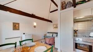A dining area with a wooden table set for two, a sofa, and a kitchen with a stove and oven. - 1 Bed in Aldeburgh (oc-al105) (Aldeburgh)