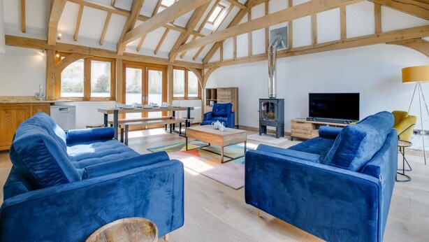 A spacious living area with a high ceiling, wooden beams, a fireplace, a television, and blue upholstered furniture. - 4 Bed in Eastbridge (oc-bricot) (East Bridge)