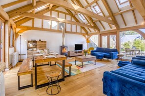 Een ruime woonkamer met een hoge plafond, houten balken, een open haard, een televisie en een blauwe bank.