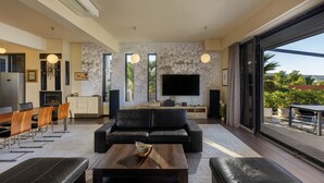 A modern living room with a black leather sofa, a wooden coffee table, and a TV mounted on the wall. There is a dining area with a wooden table and chairs, and a kitchen area with a refrigerator and a stove. - Villa Bella with heated pool (Barotul)