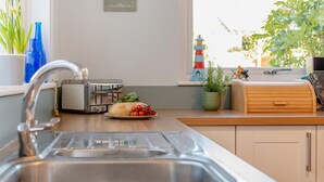 A kitchen with a stainless steel sink, a toaster, a plate with bread and tomatoes, and a window with a view of a lighthouse. - 3 Bed in Padstow (oc-p00550) (Padstow)