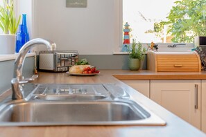 A kitchen with a stainless steel sink, a toaster, a plate with bread and tomatoes, and a window with a view of a lighthouse. - 3 Bed in Padstow (oc-p00550) (Padstow)