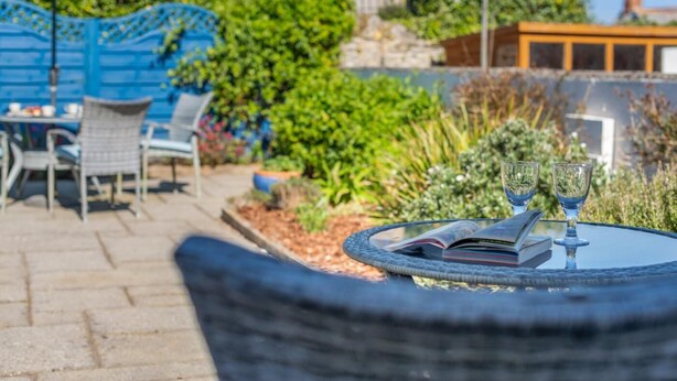 A patio area with a round table, two wine glasses, and a book. There are chairs and an umbrella in the background. - 3 Bed in Padstow (oc-p00550) (Padstow)