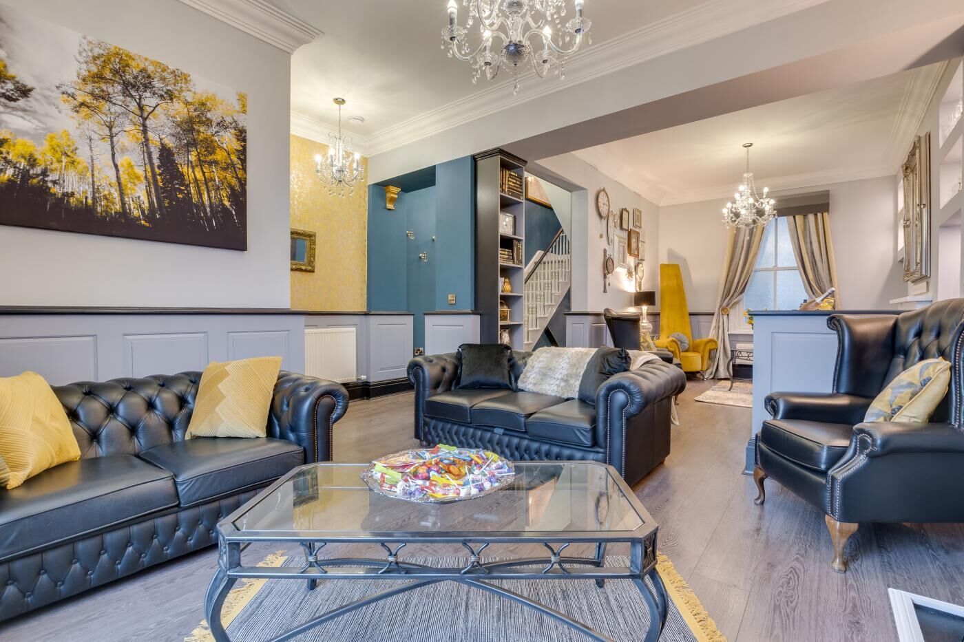 A spacious living room with a large black leather sofa, a glass coffee table, and a chandelier. There is a staircase leading to an upper level, and a painting of trees on the wall.