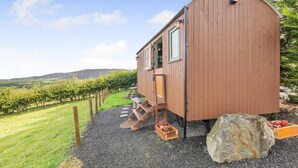 A wooden cabin with a curved roof, a front door, and a window. There is a gravel path leading to the cabin, and a large rock is visible in the foreground. - 1 Bed in Keswick (oc-f33118) (Underskiddaw)