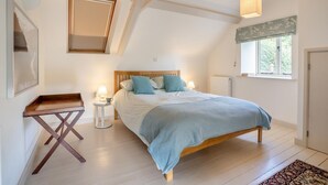 A bedroom with a wooden bed, a bedside table with a lamp, a small table with a tray, a window with a view, and a radiator.