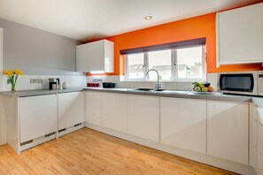A modern kitchen with white cabinets, a wooden floor, and a window with orange walls. - 3 Bed in St. Merryn (oc-p00569) (Saint Merryn)