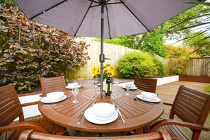 A round wooden table set for four with white plates and glasses, under a large umbrella, surrounded by wooden chairs. - 3 Bed in St. Merryn (oc-p00569) (Saint Merryn)