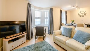 Ein Wohnzimmer mit Fernseher, einer Couch, einem Sessel und einem Essbereich mit Tisch für zwei Personen.