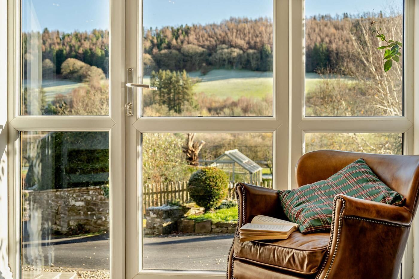 Un fauteuil en cuir avec un coussin à carreaux, placé devant une grande fenêtre à carreaux, qui offre une vue sur un paysage rural avec des arbres et un mur de pierres.