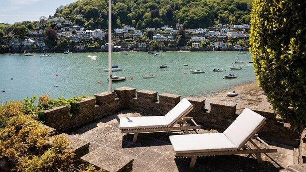 Une terrasse en bord de mer, avec des murs en pierre et des fauteuils confortables, offrant une vue sur un port où sont amarrés des bateaux et sur une colline parsemée de maisons.