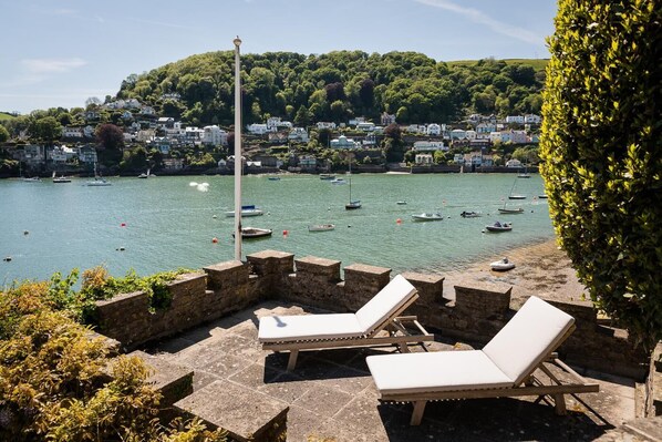 A seaside patio with stone walls and lounge chairs, overlooking a harbor with boats and a hillside of houses.