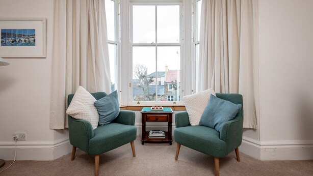 Ein Zimmer mit zwei grünen Sesseln, einem kleinen Holztisch, einem gerahmten Bild an der Wand und einem Erkerfenster mit Vorhängen.