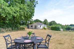 An outdoor dining area with a round table and chairs, a vase with flowers, and a house in the background.