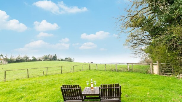 Une table de pique-nique avec deux bancs, située dans un champ herbeux, avec une clôture et des arbres en arrière-plan.