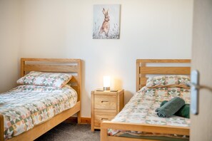 A room with two wooden beds, each with a patterned bedspread and pillows, a bedside table with a lamp, and a wall-mounted painting.