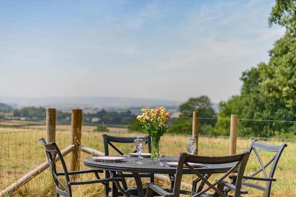 An outdoor dining area with a table and chairs, a vase of flowers, and a view of the countryside.