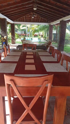 Un espacio de comedor al aire libre cubierto, con una mesa larga y sillas de madera, rodeado de follaje y una piscina.