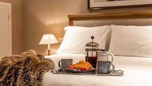 Una cama impecablemente tendida con sábanas blancas, un plaid de piel, una bandeja con un croissant, una cafetera y una taza.