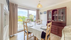A dining area with a table set for four, chairs, a cabinet with glassware, and a view through a large window. - 3 Bed in Paignton (oc-bc1890) (Paignton)