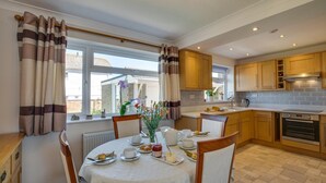 A modern kitchen with wooden cabinets, a dining table set for breakfast, and a view of the outside. - 3 Bed in Paignton (oc-puffin) (Paignton)