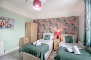 A bedroom with two beds, a wardrobe, a nightstand with a lamp, and a floral wallpaper. - 6 Bed in Llandudno (oc-s33417) (Llandudno)