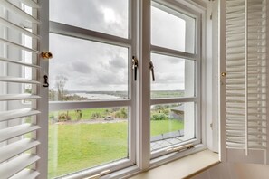 Ein Zimmer mit großem Fenster, weißen Jalousien und Blick auf eine grüne Landschaft.
