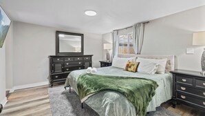 Une chambre à coucher comprenant un lit, une commode, une table de chevet, un miroir et une fenêtre avec des rideaux.