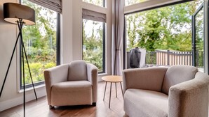 A modern living room with a sofa, armchair, and floor lamp, large windows with curtains, and a view of greenery outside. - 4 Bed in Coleford (oc-t33301) (Coleford)