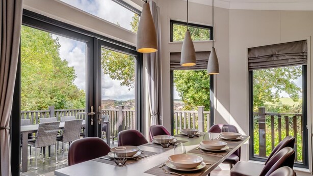 A dining area with a long table set for six, large windows offering a view of greenery and hills, and pendant lighting above. - 4 Bed in Coleford (oc-t33301) (Coleford)