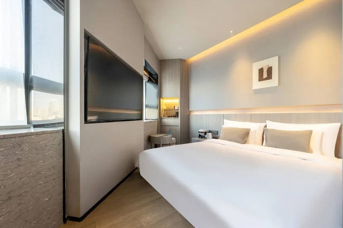 A modern hotel room with a large bed, a flat-screen TV mounted on the wall, a small bedside table, and a view of the city through the window.