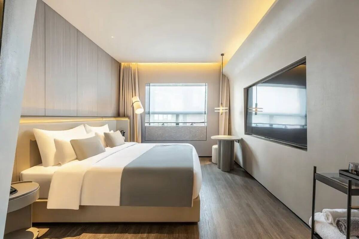 A modern hotel room with a large bed, a bedside table, a round table, and a large window with a view.