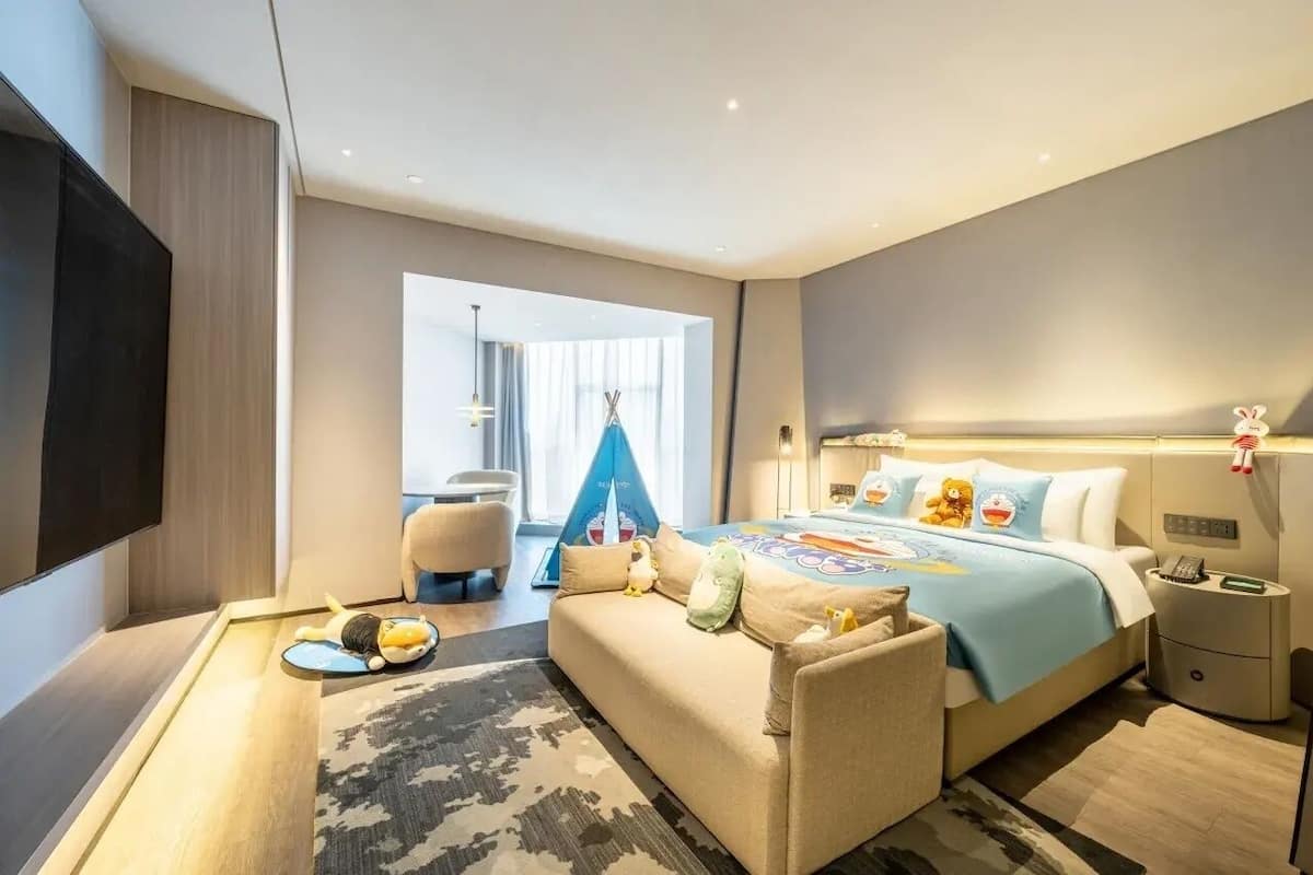 A modern hotel room with a sofa, a bed with plush pillows, a bedside table with a lamp, and a large window with curtains.