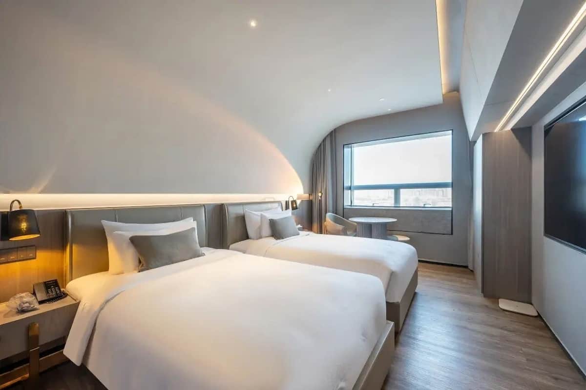 A modern hotel room with two beds, a desk, a chair, a small table, and a large window offering a city view.