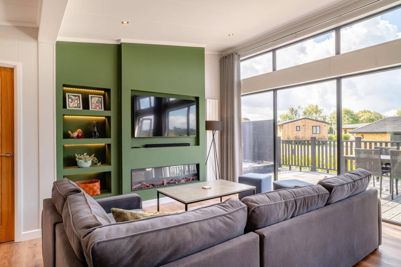 Een moderne woonkamer met een groene accentmuur, een flatscreen televisie, een open haard en een groot raam met uitzicht op een terras.