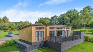 A modern wooden cabin with a deck, large windows, and a car parked nearby. - 2 Bed in Forest Of Dean (oc-t33299) (Coleford)