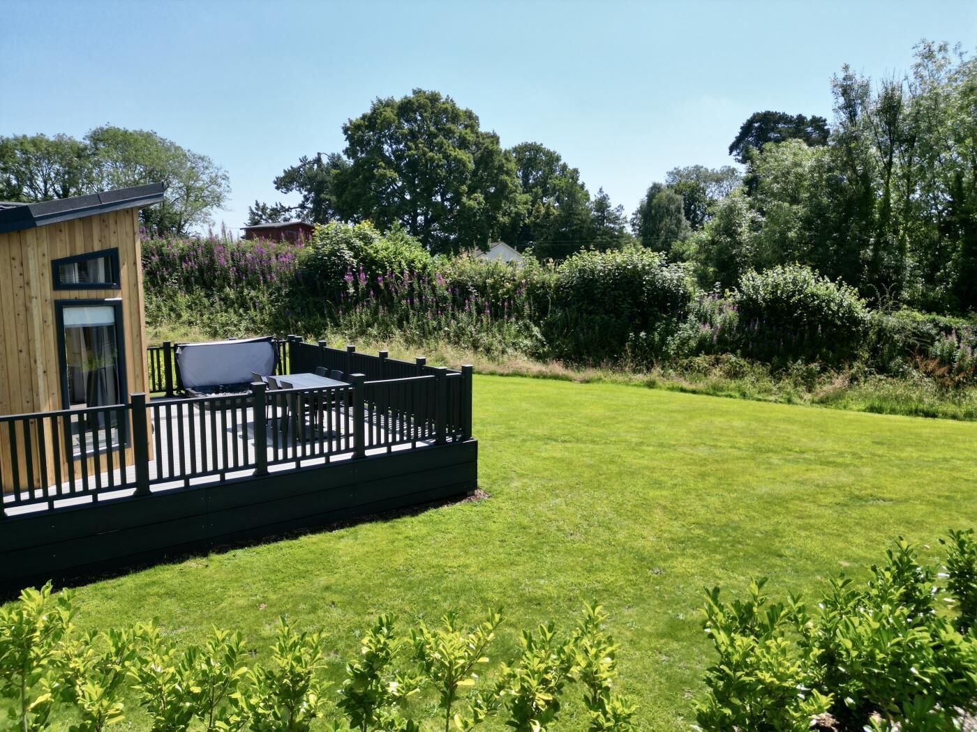 Een houten chalet met een terras, omringd door een verzorgd gazon en weelderige struiken.