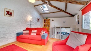 A cozy living room with a red sofa, wooden beams on the ceiling, a skylight, and a view of the kitchen through an open door. - 1 Bed in Totnes (oc-bcbove) (Broadhempston)