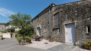 A stone building with a white door, a small window, and a paved courtyard with a table and chairs. - 2 Bed in Totnes (oc-bcteig) (Broadhempston)