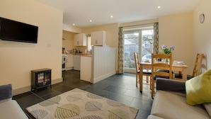 A modern living room with a sofa, a TV mounted on the wall, and a dining area with a table and chairs. - 2 Bed in Totnes (oc-bcteig) (Broadhempston)