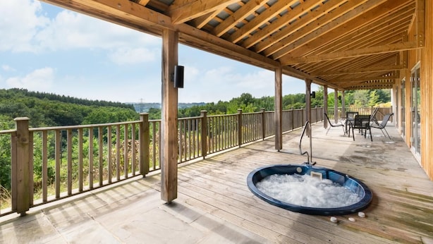 A covered outdoor deck with a hot tub, wooden railing, and a view of a forested landscape. - 2 Bed in Totnes (oc-bcteig) (Broadhempston)