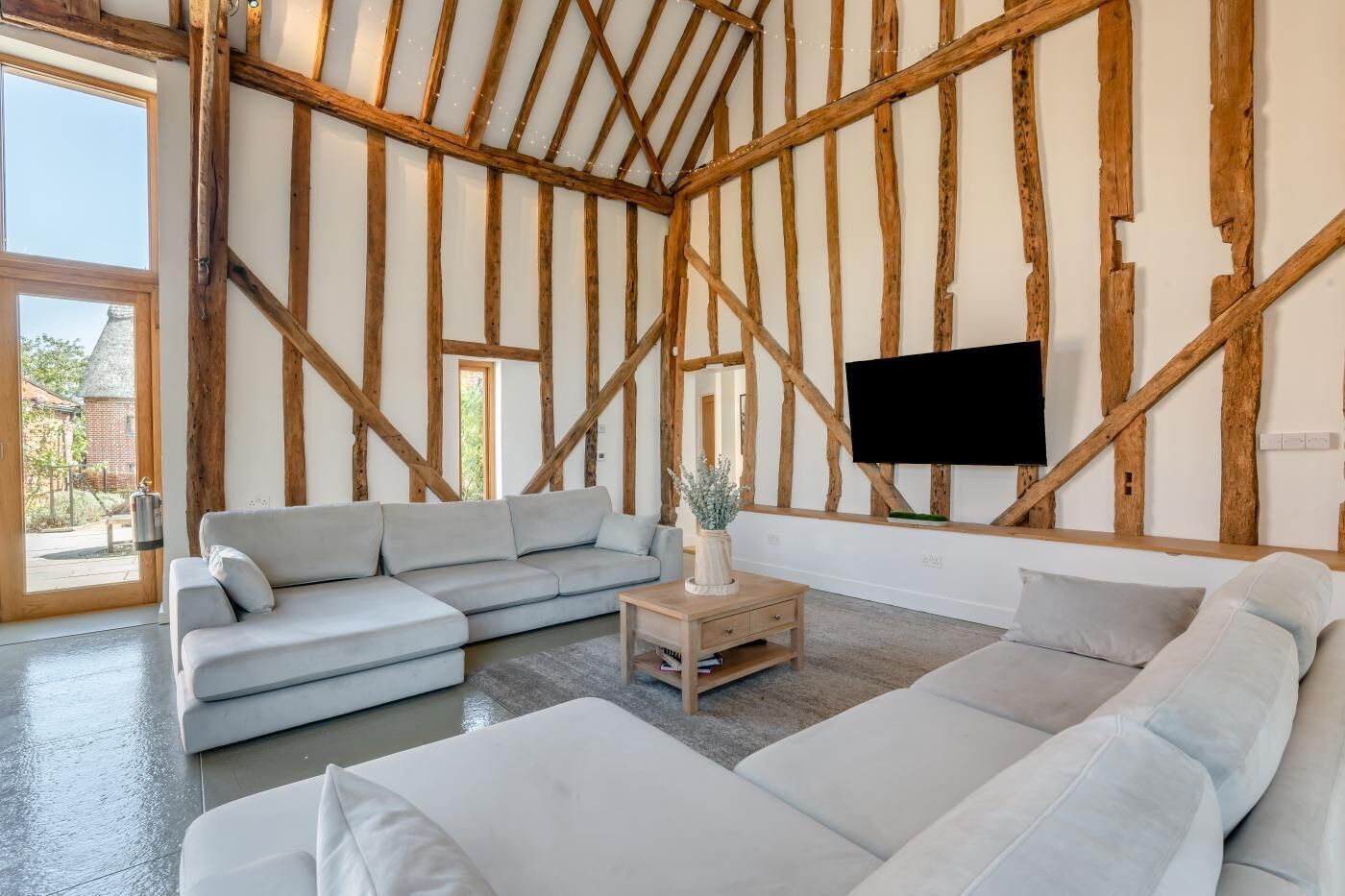 Een moderne woonkamer met zichtbare houten balken, een grijze modulaire bank, een klein houten bijzettafeltje en een flatscreen televisie aan de wand.