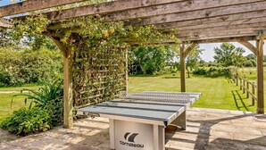Ein Tischtennis-Tisch im Freien unter einer Holz-Pergola mit Garten im Hintergrund.