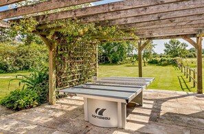 Ein Tischtennis-Tisch im Freien unter einer Holz-Pergola mit Garten im Hintergrund.