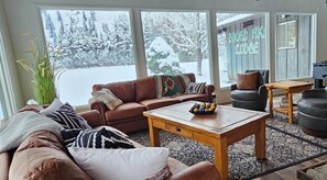 A cozy living room with a brown leather sofa, a wooden coffee table, and a view of a snowy landscape through large windows.
